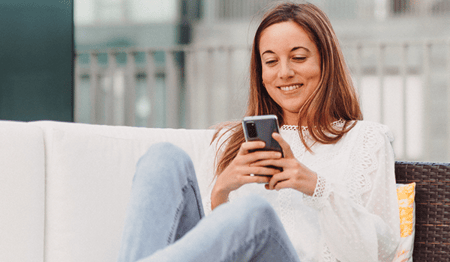 Eine Frau sitzt lächelnd auf einer Couch und schaut auf ihr Smartphone.